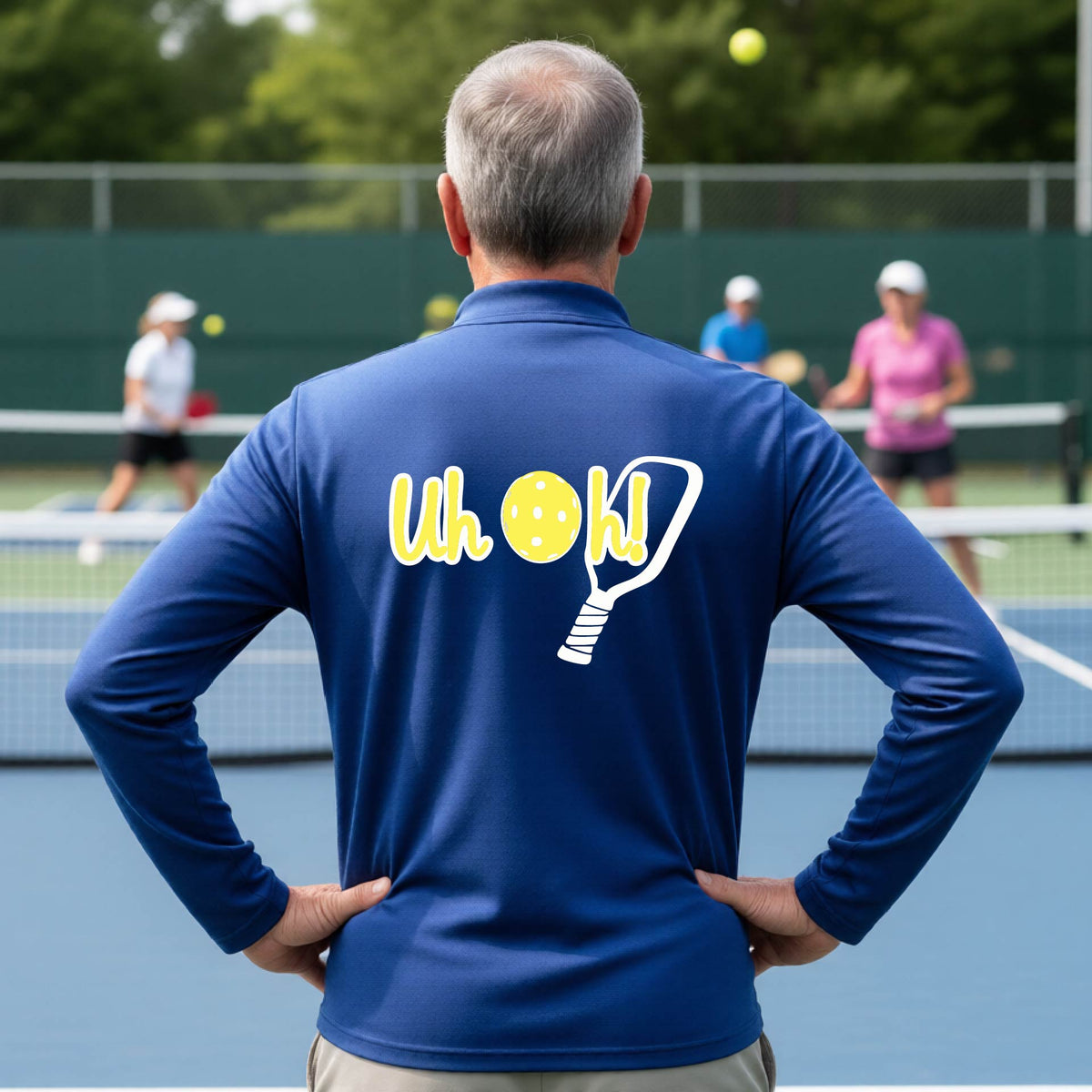 A man seen from behind on a pickleball court wearing a royal blue quarter-zip shirt with a white and yellow graphic of the words “Uh Oh!” with a yellow pickleball replacing the letter O, over the top of a pickleball paddle, on the back of the shirt.