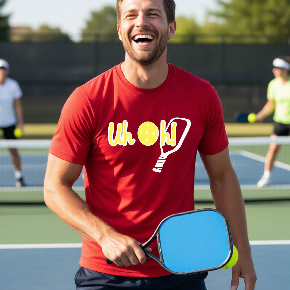 A man on a pickleball court wearing a red short-sleeve t-shirt with a white and yellow graphic of the words “Uh Oh!” with a yellow pickleball replacing the letter O on the front of the shirt, over the top of a pickleball paddle.