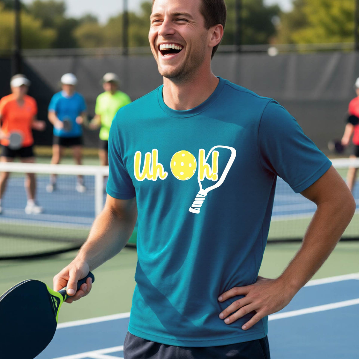 A man on a pickleball court wearing a teal short-sleeve t-shirt with a white and yellow graphic of the words “Uh Oh!” with a yellow pickleball replacing the letter O on the front of the shirt, over the top of a pickleball paddle.