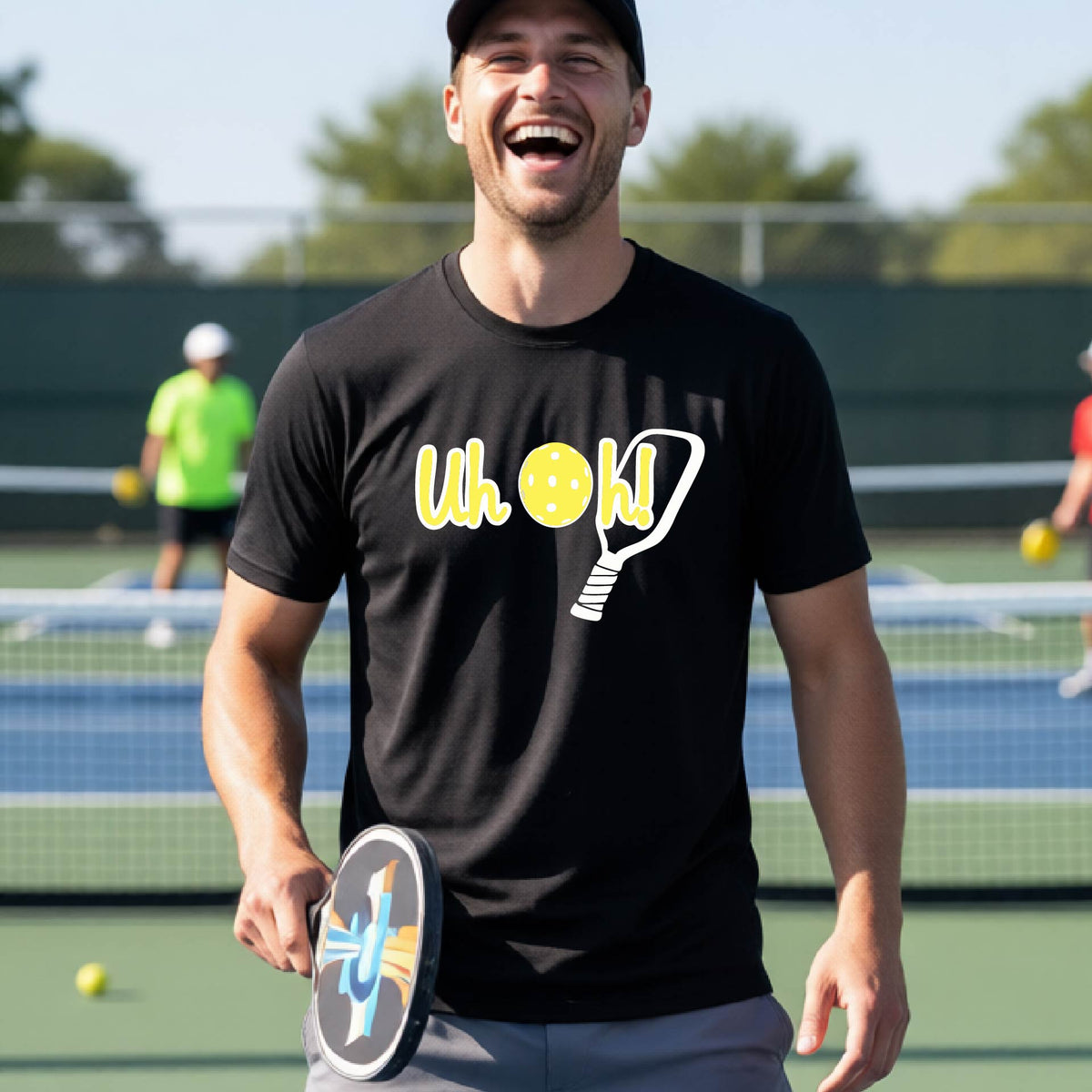 A man on a pickleball court wearing a black short-sleeve t-shirt with a white and yellow graphic of the words “Uh Oh!” with a yellow pickleball replacing the letter O on the front of the shirt, over the top of a pickleball paddle.