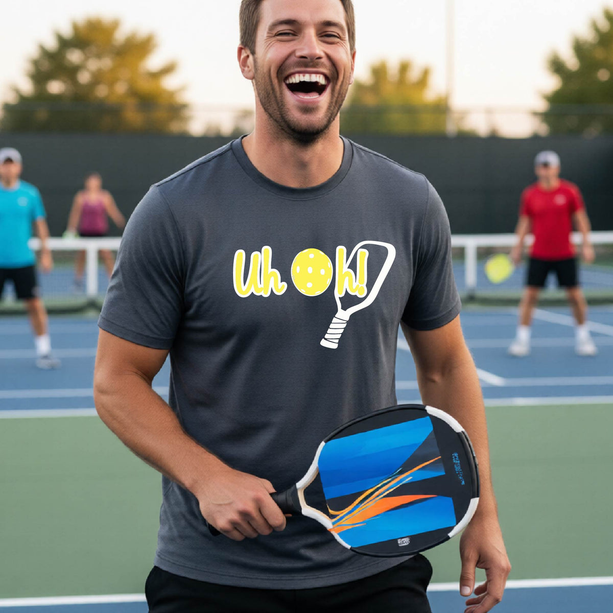 A man on a pickleball court wearing a gray short-sleeve t-shirt with a white and yellow graphic of the words “Uh Oh!” with a yellow pickleball replacing the letter O on the front of the shirt, over the top of a pickleball paddle.