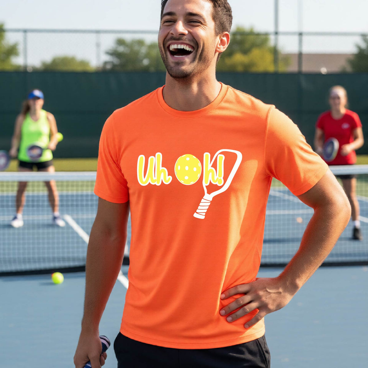 A man on a pickleball court wearing an orange short-sleeve t-shirt with a white and yellow graphic of the words “Uh Oh!” with a yellow pickleball replacing the letter O on the front of the shirt, over the top of a pickleball paddle.