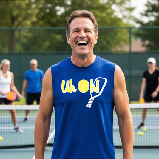 A man on a pickleball court wearing a royal  blue sleeveless t-shirt with a white and yellow graphic of the words “Uh Oh!” with a yellow pickleball replacing the letter O on the front of the shirt, over the top of a pickleball paddle.