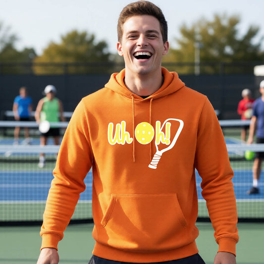 A man on a pickleball court wearing an orange hoodie pullover with a white and yellow graphic of the words “Uh Oh!” with a yellow pickleball replacing the letter O over the top of a pickleball paddle, on the front of the hoodie.