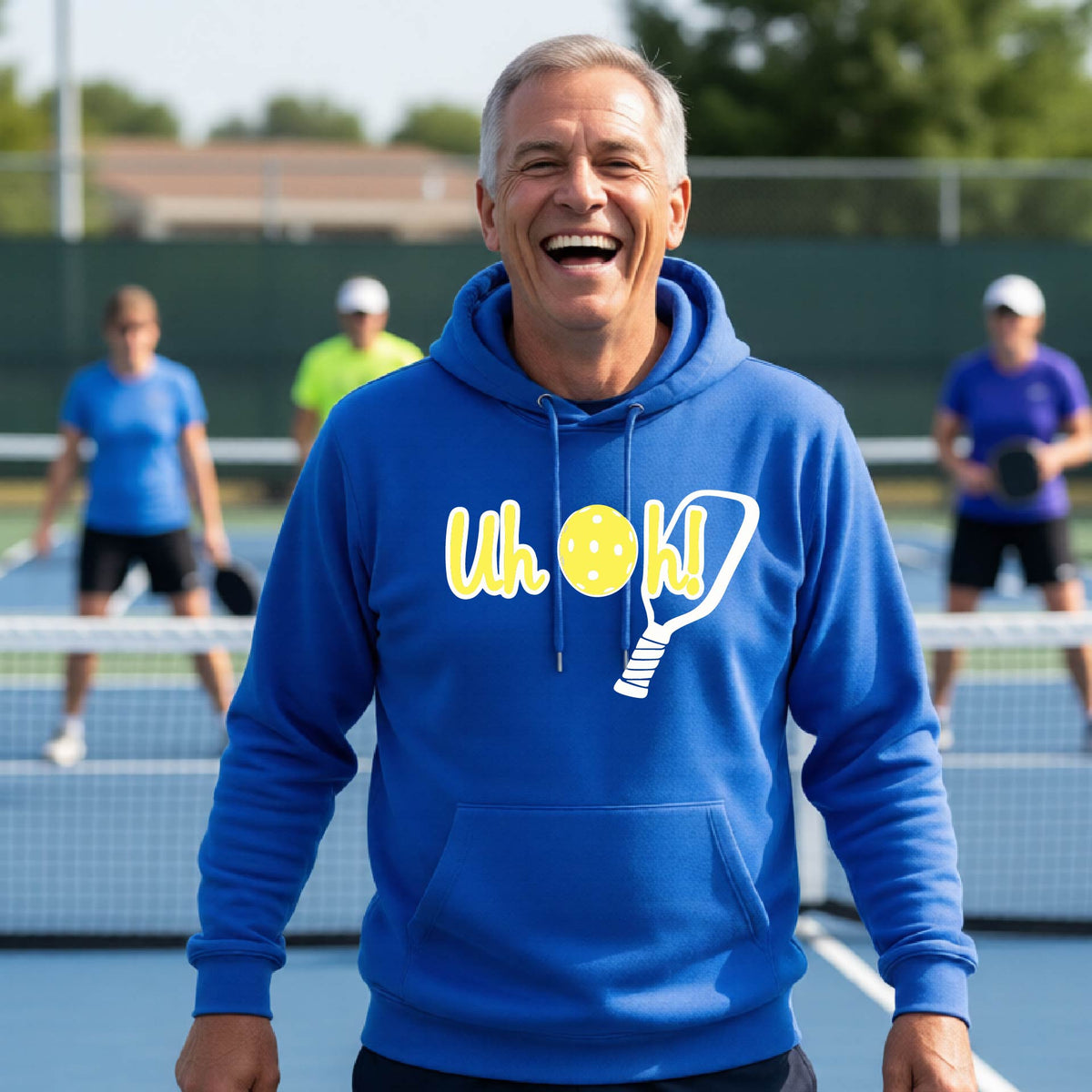 A man on a pickleball court wearing a royal blue hoodie pullover with a white and yellow graphic of the words “Uh Oh!” with a yellow pickleball replacing the letter O over the top of a pickleball paddle, on the front of the hoodie.