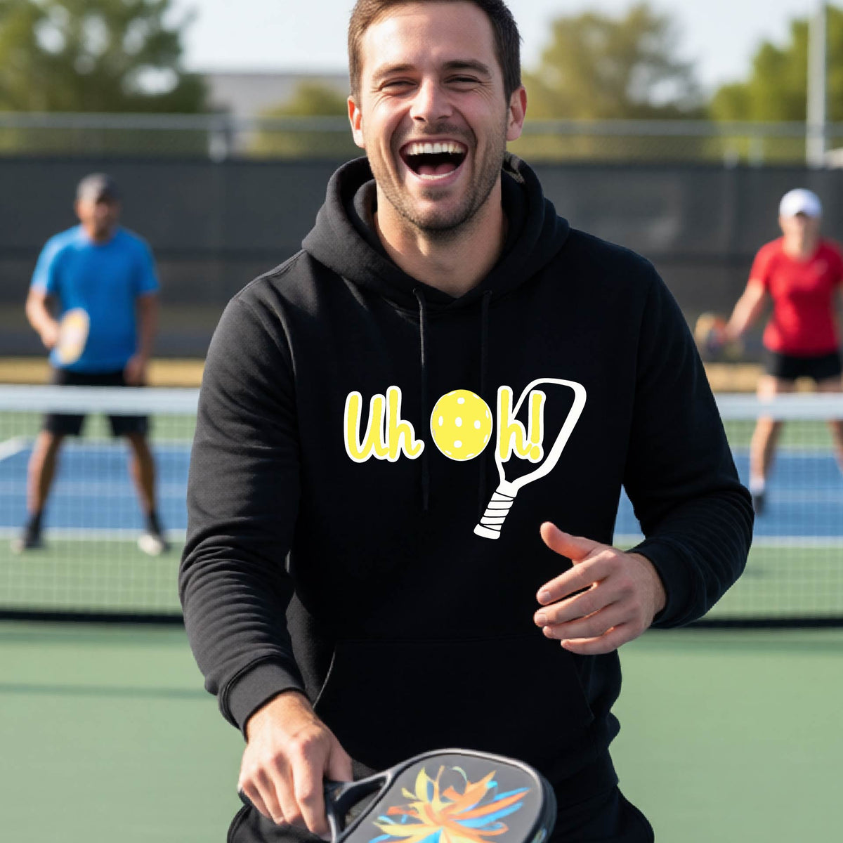 A man on a pickleball court wearing a black hoodie pullover with a white and yellow graphic of the words “Uh Oh!” with a yellow pickleball replacing the letter O over the top of a pickleball paddle, on the front of the hoodie.