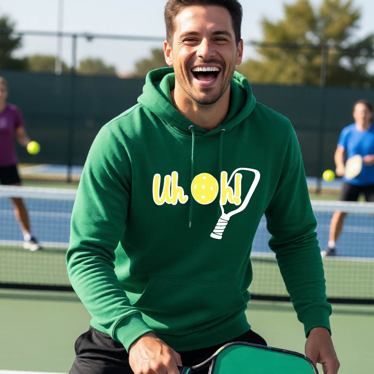 A man on a pickleball court wearing a green hoodie pullover with a white and yellow graphic of the words “Uh Oh!” with a yellow pickleball replacing the letter O over the top of a pickleball paddle, on the front of the hoodie.