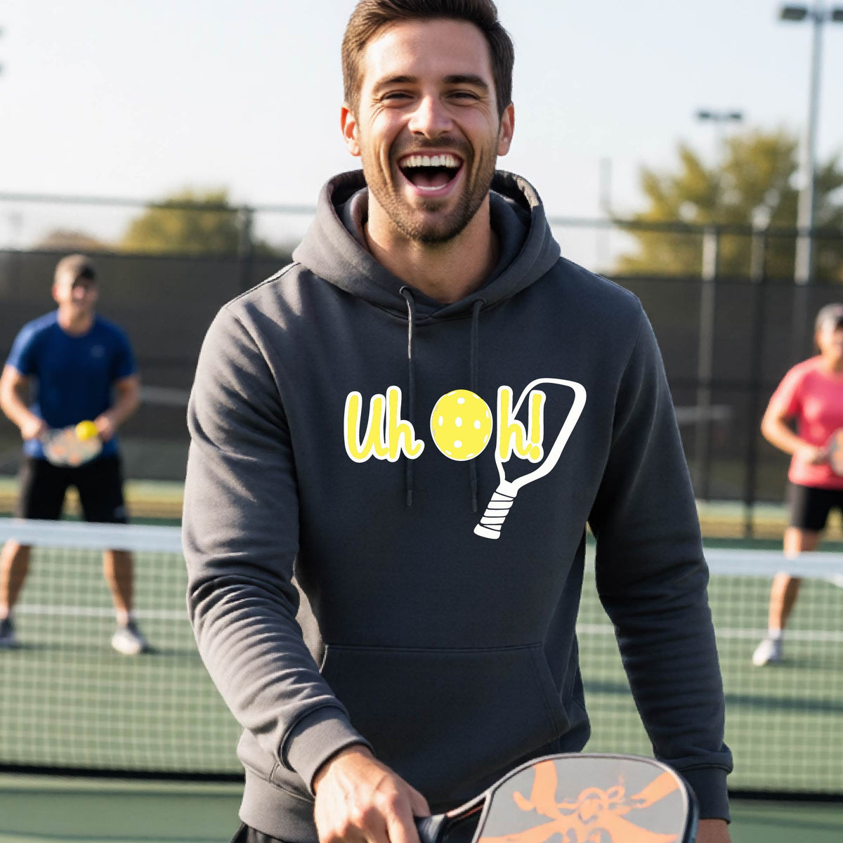 A man on a pickleball court wearing a gray hoodie pullover with a white and yellow graphic of the words “Uh Oh!” with a yellow pickleball replacing the letter O over the top of a pickleball paddle, on the front of the hoodie.