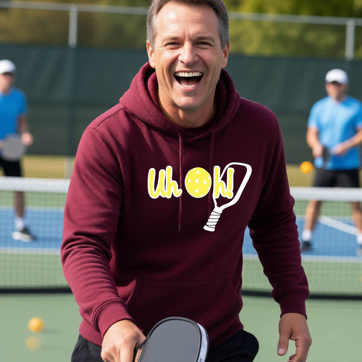 A man on a pickleball court wearing a maroon hoodie pullover with a white and yellow graphic of the words “Uh Oh!” with a yellow pickleball replacing the letter O over the top of a pickleball paddle, on the front of the hoodie.