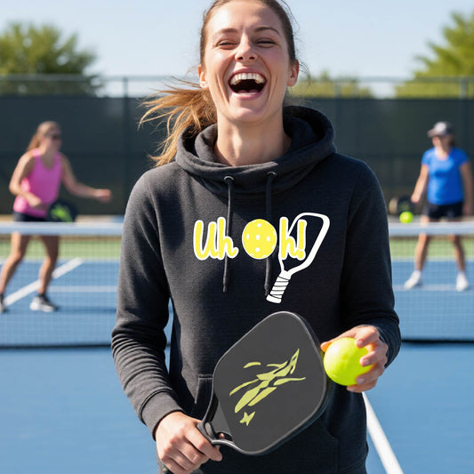 A woman on a pickleball court wearing a black cowl-neck hoodie pullover with a white and yellow graphic of the words “Uh Oh!” with a yellow pickleball replacing the letter O over the top of a pickleball paddle, on the front of the hoodie.

