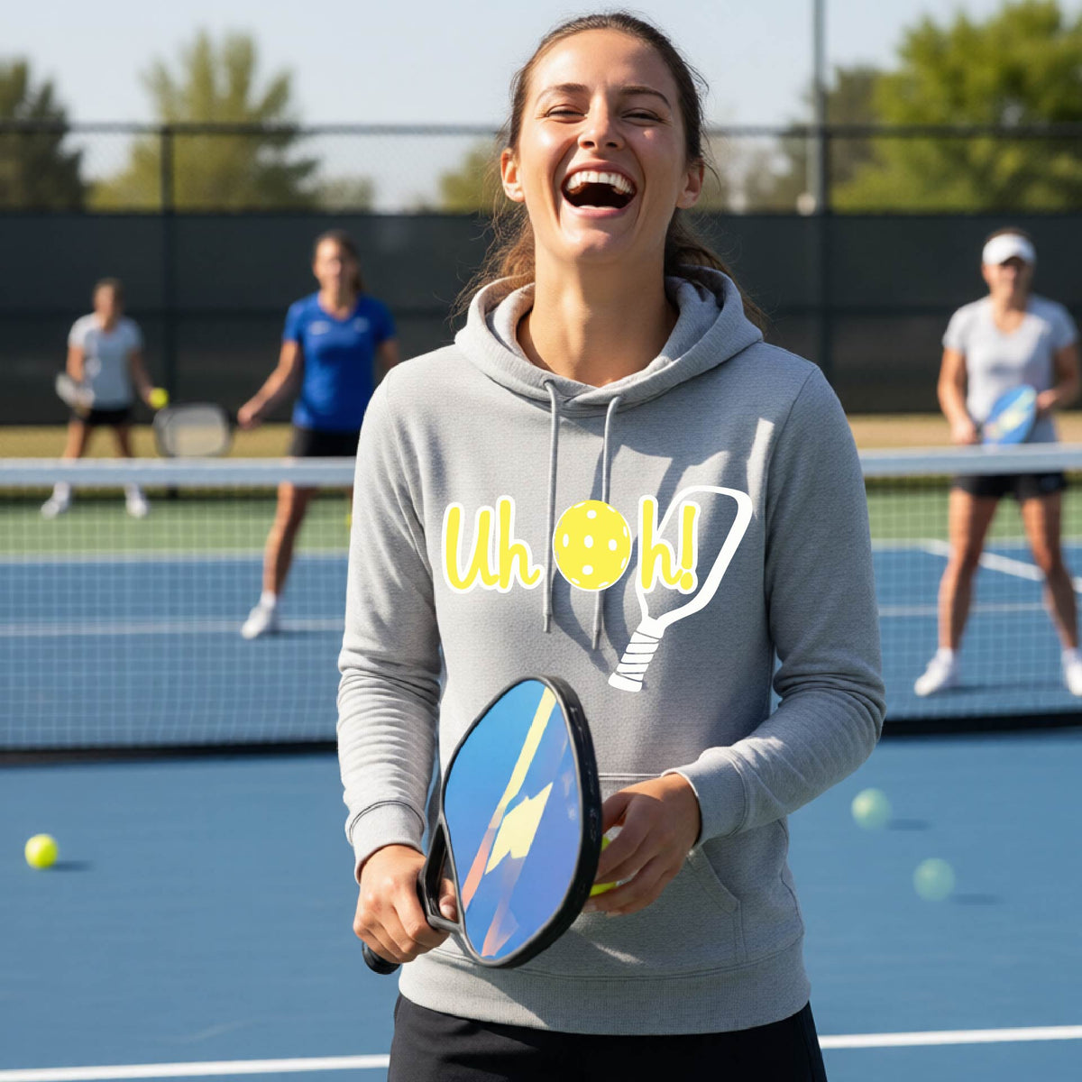 A woman on a pickleball court wearing a light gray hoodie pullover with a white and yellow graphic of the words “Uh Oh!” with a yellow pickleball replacing the letter O over the top of a pickleball paddle, on the front of the hoodie.

