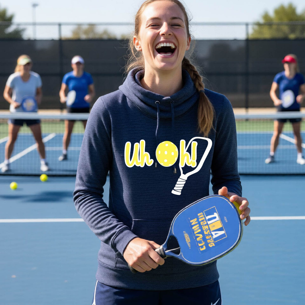 A woman on a pickleball court wearing a navy blue hoodie pullover with a white and yellow graphic of the words “Uh Oh!” with a yellow pickleball replacing the letter O over the top of a pickleball paddle, on the front of the hoodie.

