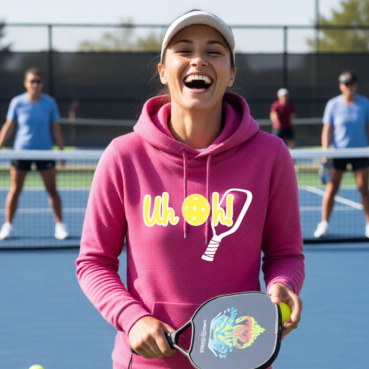 A woman on a pickleball court wearing a pink hoodie pullover with a white and yellow graphic of the words “Uh Oh!” with a yellow pickleball replacing the letter O over the top of a pickleball paddle, on the front of the hoodie.

