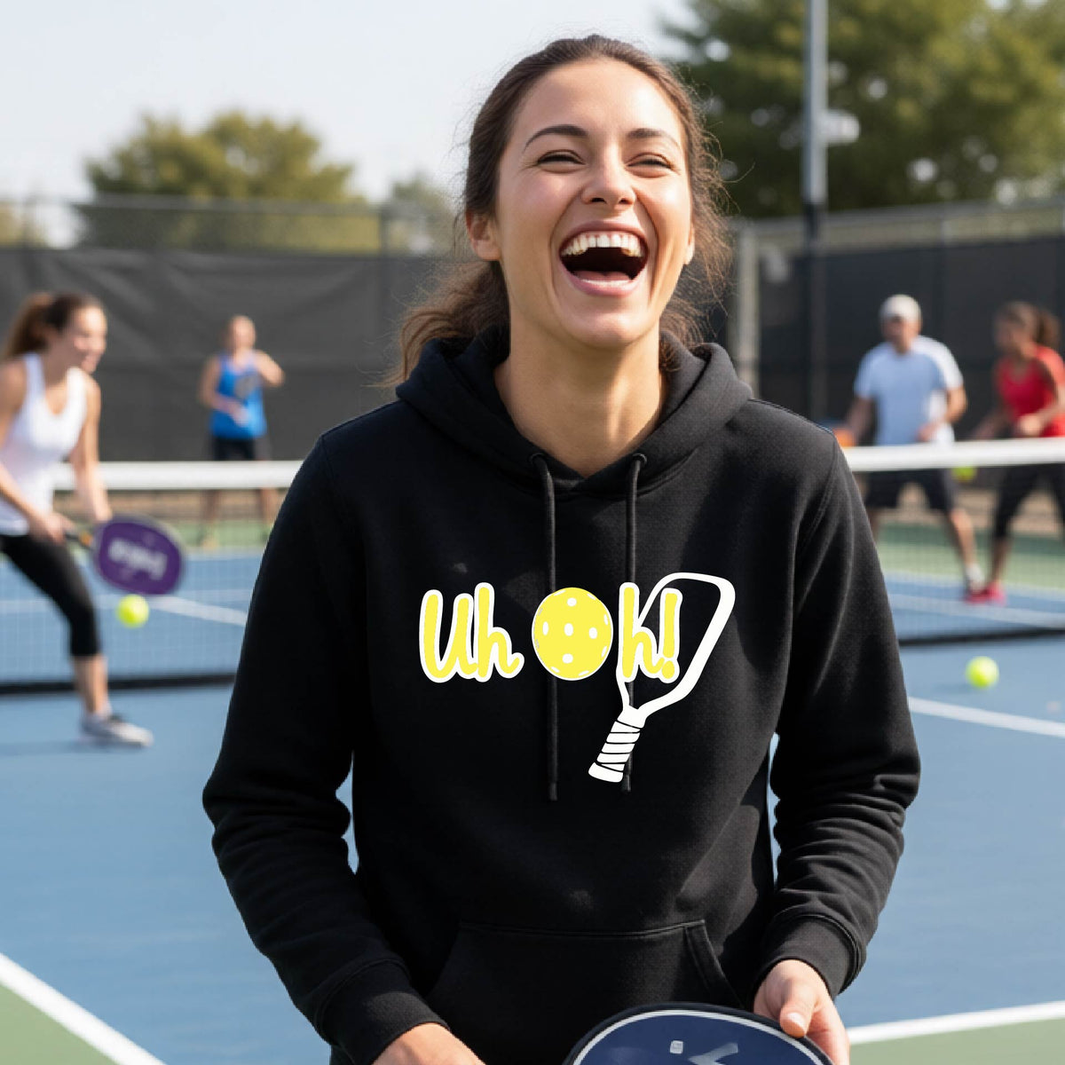 A woman on a pickleball court wearing a black hoodie pullover with a white and yellow graphic of the words “Uh Oh!” with a yellow pickleball replacing the letter O over the top of a pickleball paddle, on the front of the hoodie.