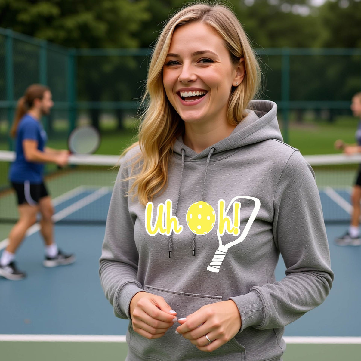 A woman on a pickleball court wearing a gray hoodie pullover with a white and yellow graphic of the words “Uh Oh!” with a yellow pickleball replacing the letter O over the top of a pickleball paddle, on the front of the hoodie.