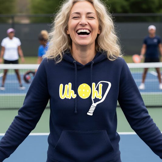 A woman on a pickleball court wearing a navy blue hoodie pullover with a white and yellow graphic of the words “Uh Oh!” with a yellow pickleball replacing the letter O over the top of a pickleball paddle, on the front of the hoodie.