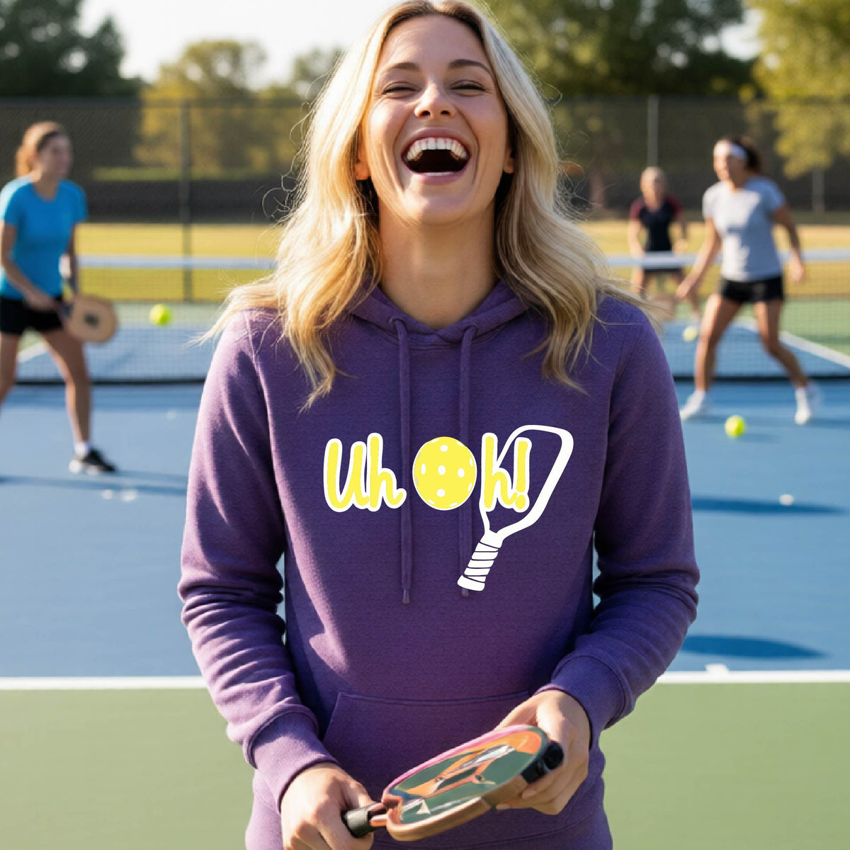 A woman on a pickleball court wearing a purple hoodie pullover with a white and yellow graphic of the words “Uh Oh!” with a yellow pickleball replacing the letter O over the top of a pickleball paddle, on the front of the hoodie.