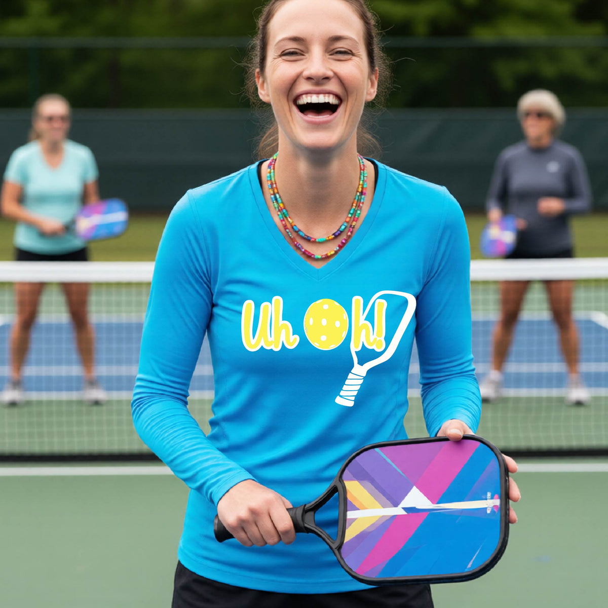 A woman on a pickleball court wearing a bright blue long-sleeve t-shirt with a white and yellow graphic of the words “Uh Oh!” with a yellow pickleball replacing the letter O on the front of the shirt, over the top of a pickleball paddle.