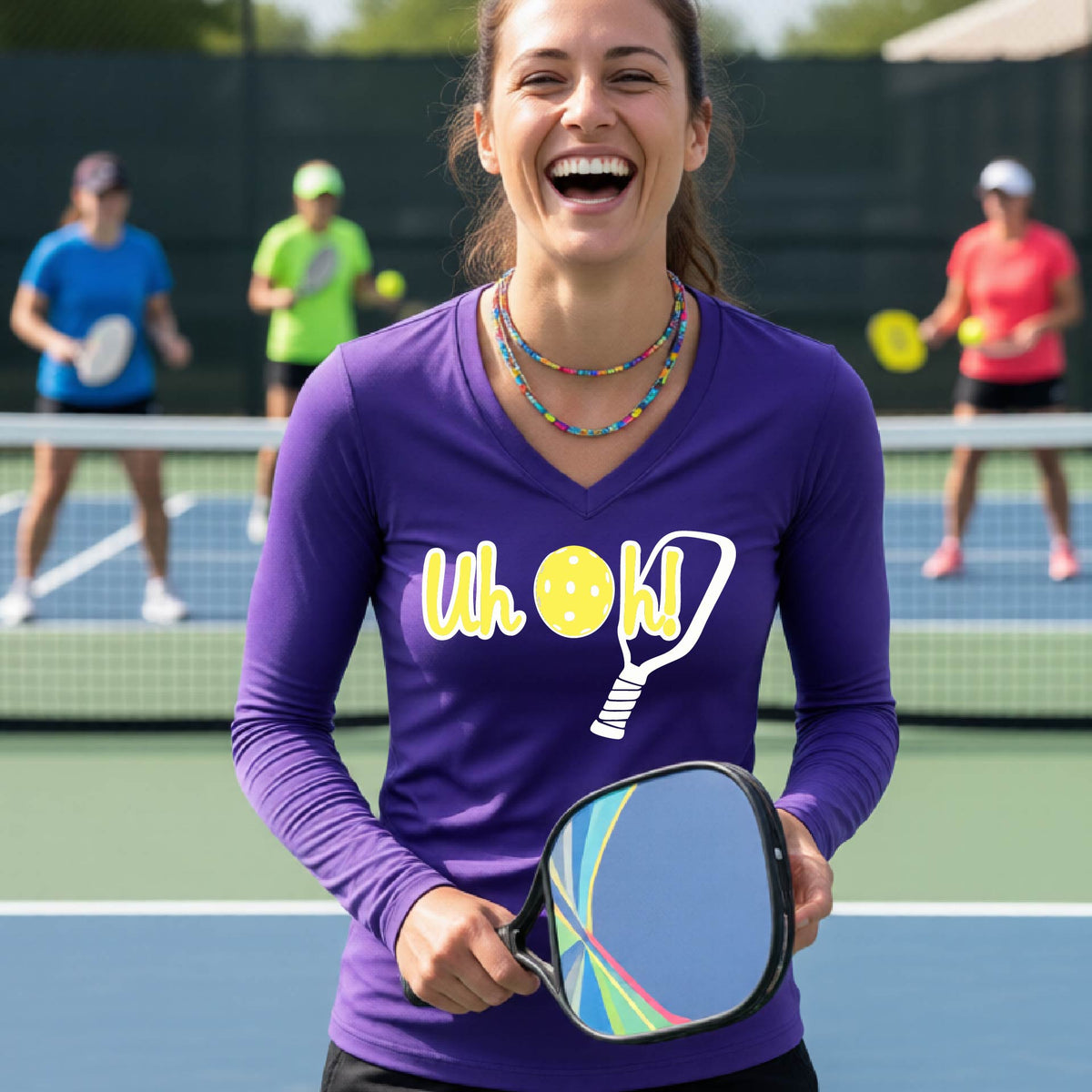 A woman on a pickleball court wearing a purple long-sleeve t-shirt with a white and yellow graphic of the words “Uh Oh!” with a yellow pickleball replacing the letter O on the front of the shirt, over the top of a pickleball paddle.
