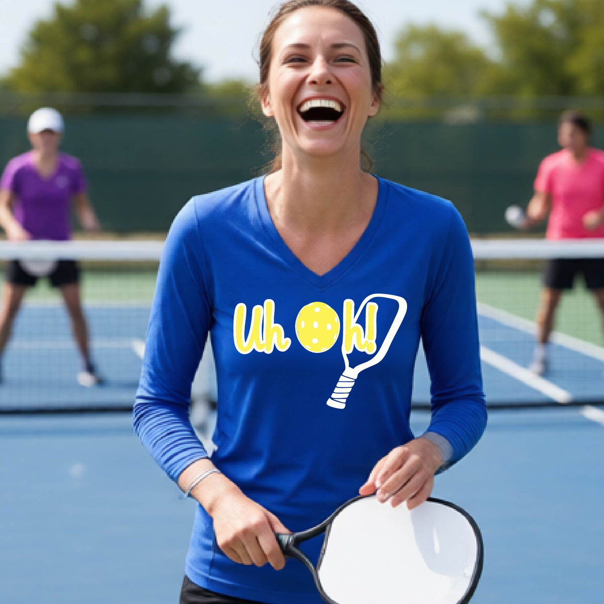 A woman on a pickleball court wearing a royal blue long-sleeve t-shirt with a white and yellow graphic of the words “Uh Oh!” with a yellow pickleball replacing the letter O on the front of the shirt, over the top of a pickleball paddle.
