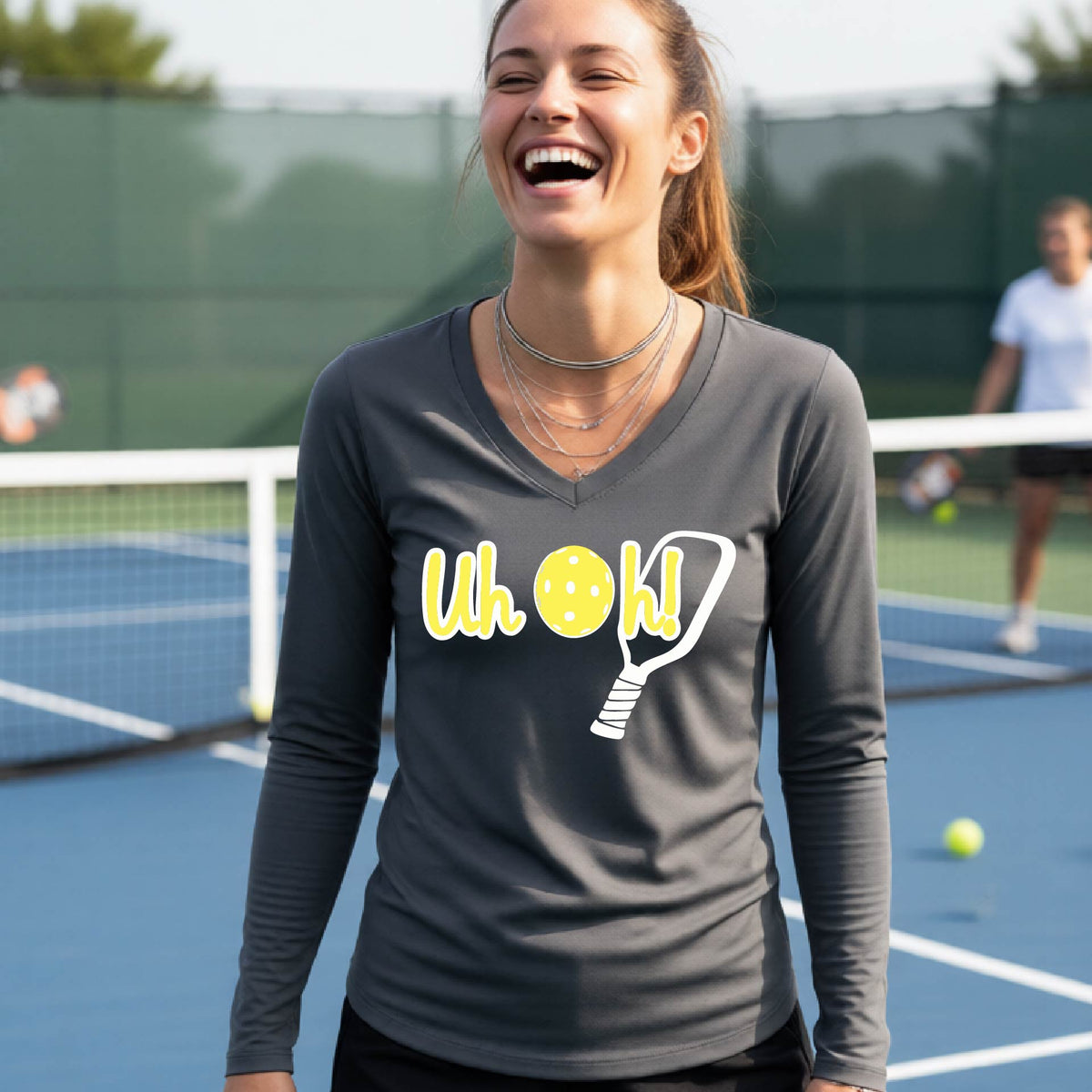 A woman on a pickleball court wearing a gray long-sleeve t-shirt with a white and yellow graphic of the words “Uh Oh!” with a yellow pickleball replacing the letter O on the front of the shirt, over the top of a pickleball paddle.