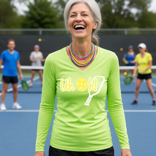 A woman on a pickleball court wearing a lime green long-sleeve t-shirt with a white and yellow graphic of the words “Uh Oh!” with a yellow pickleball replacing the letter O on the front of the shirt, over the top of a pickleball paddle.