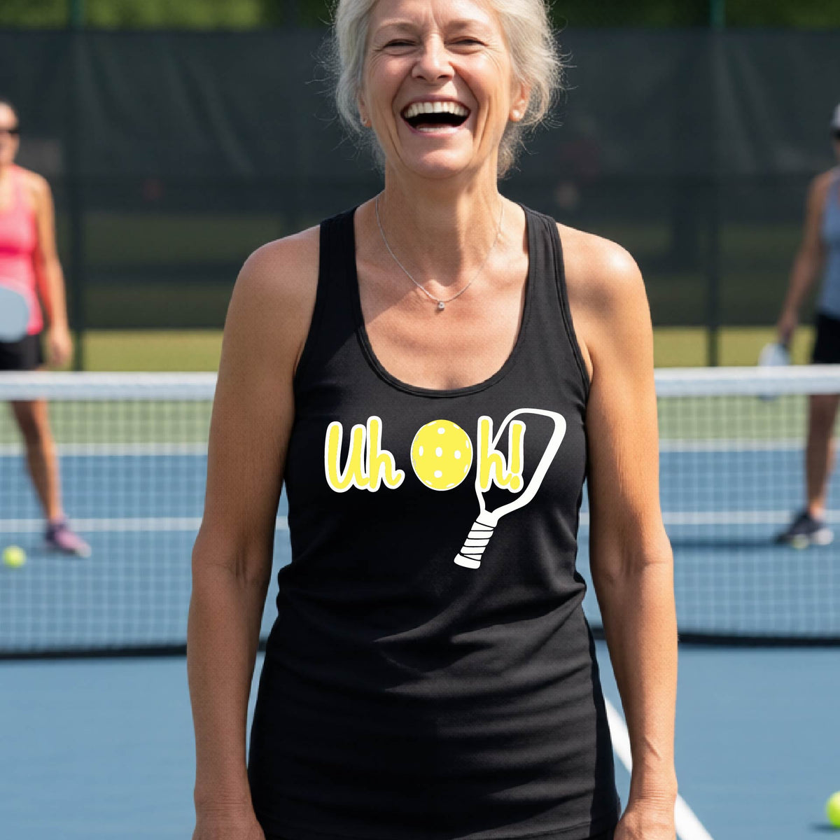 A woman on a pickleball court wearing a black racer-back tank top with a white and yellow graphic of the words “Uh Oh!” with a yellow pickleball replacing the letter O over the top of a pickleball paddle, on the front of the hoodie.