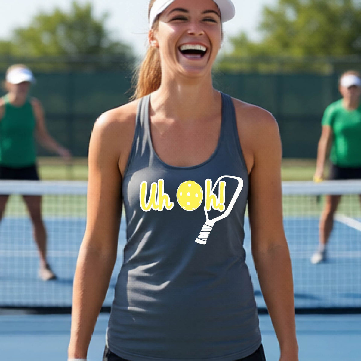 A woman on a pickleball court wearing a dark gray racer-back tank top with a white and yellow graphic of the words “Uh Oh!” with a yellow pickleball replacing the letter O over the top of a pickleball paddle, on the front of the hoodie.