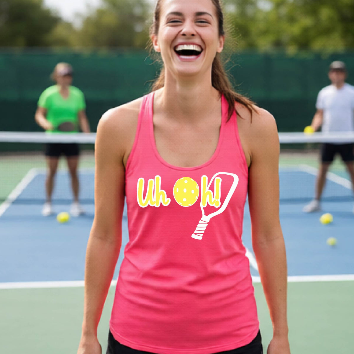 A woman on a pickleball court wearing a coral pink racer-back tank top with a white and yellow graphic of the words “Uh Oh!” with a yellow pickleball replacing the letter O over the top of a pickleball paddle, on the front of the hoodie.