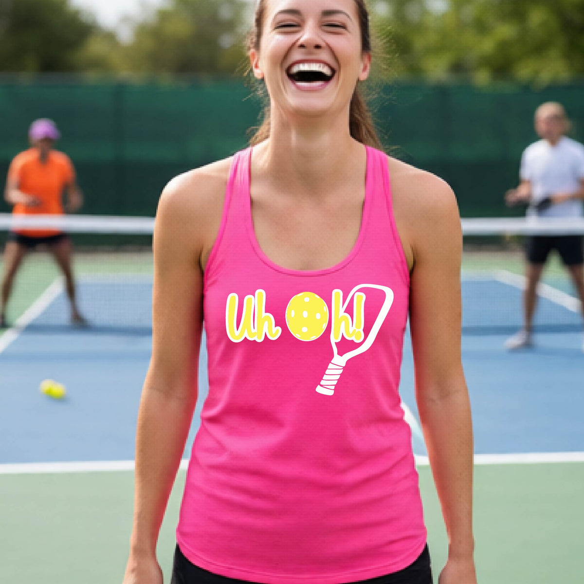 A woman on a pickleball court wearing a pink racer-back tank top with a white and yellow graphic of the words “Uh Oh!” with a yellow pickleball replacing the letter O over the top of a pickleball paddle, on the front of the hoodie.
