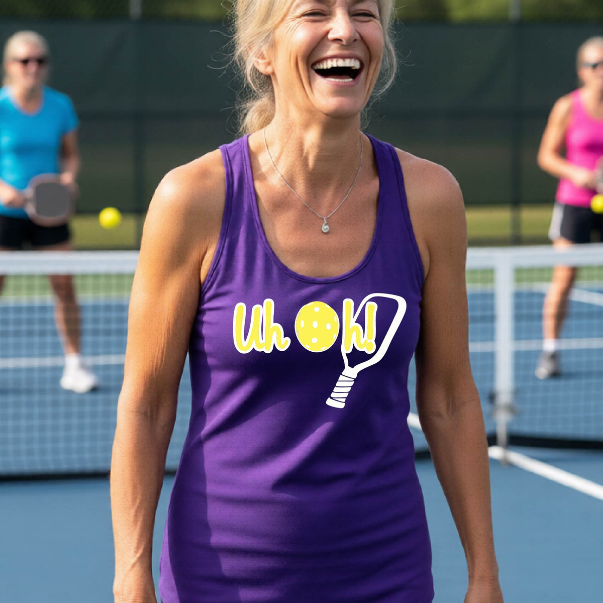 A woman on a pickleball court wearing a purple racer-back tank top with a white and yellow graphic of the words “Uh Oh!” with a yellow pickleball replacing the letter O over the top of a pickleball paddle, on the front of the hoodie.


