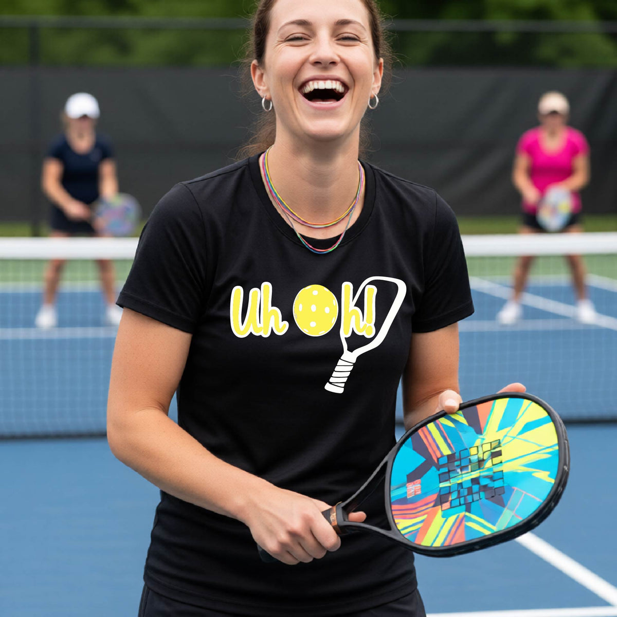 A woman on a pickleball court wearing a black short-sleeve t-shirt with a white and yellow graphic of the words “Uh Oh!” with a yellow pickleball replacing the letter O on the front of the shirt, over the top of a pickleball paddle.