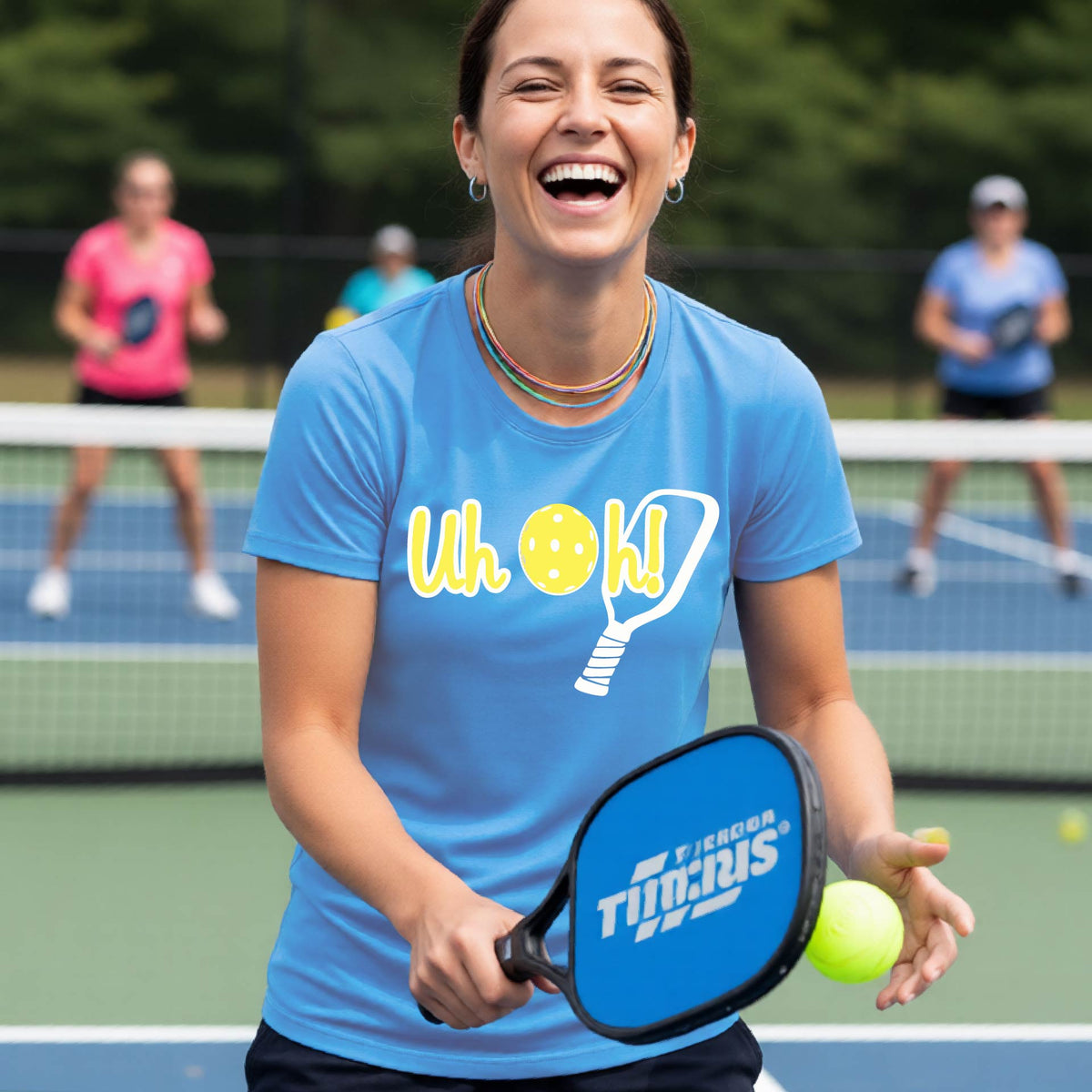 A woman on a pickleball court wearing a blue short-sleeve t-shirt with a white and yellow graphic of the words “Uh Oh!” with a yellow pickleball replacing the letter O on the front of the shirt, over the top of a pickleball paddle.