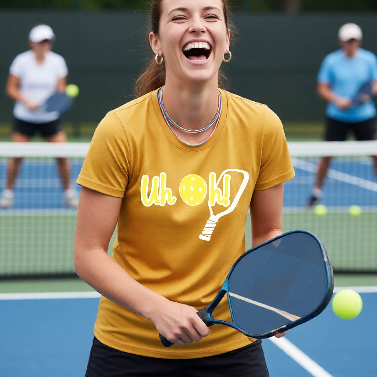 A woman on a pickleball court wearing a mustard yellow gold short-sleeve t-shirt with a white and yellow graphic of the words “Uh Oh!” with a yellow pickleball replacing the letter O on the front of the shirt, over the top of a pickleball paddle.