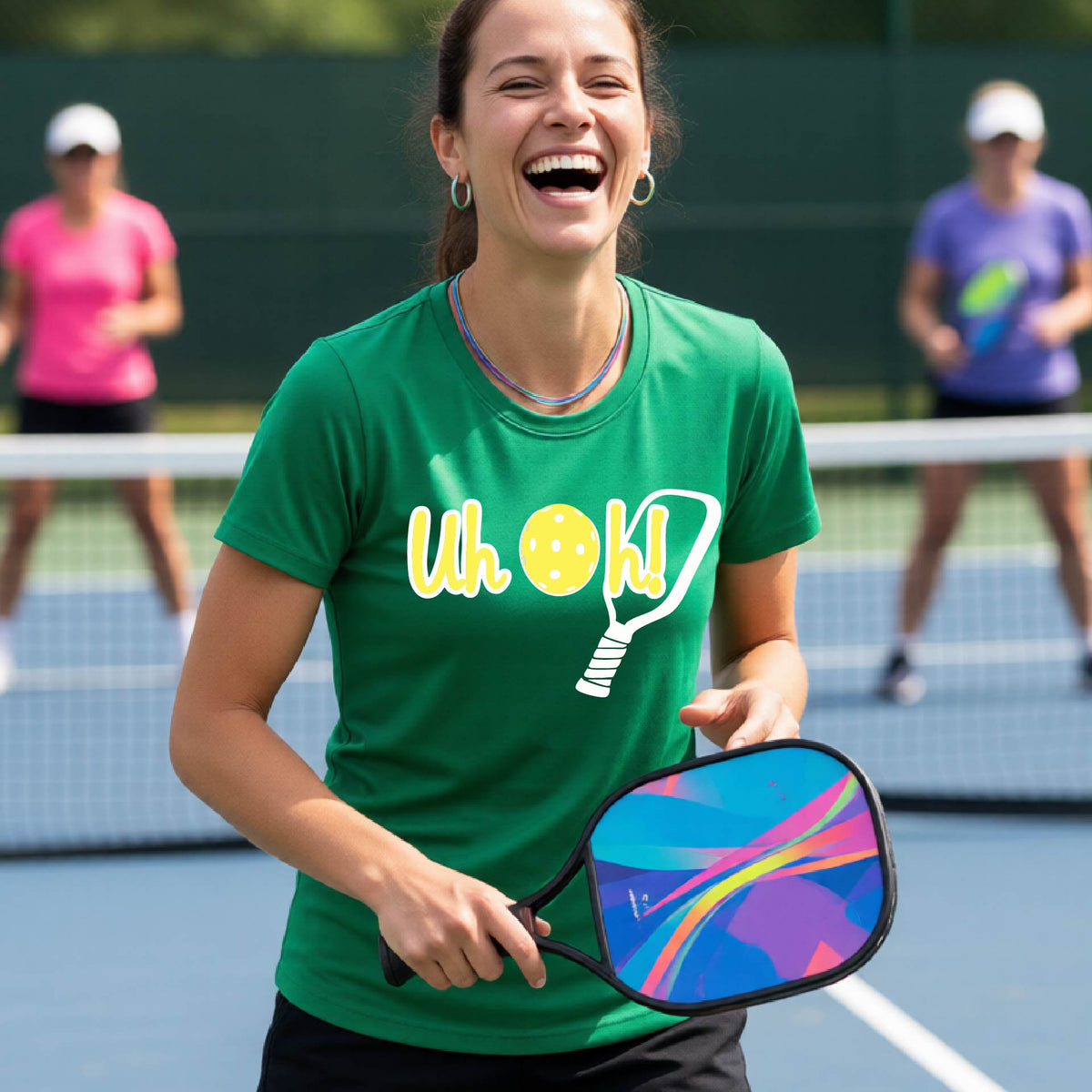 A woman on a pickleball court wearing a green short-sleeve t-shirt with a white and yellow graphic of the words “Uh Oh!” with a yellow pickleball replacing the letter O on the front of the shirt, over the top of a pickleball paddle.