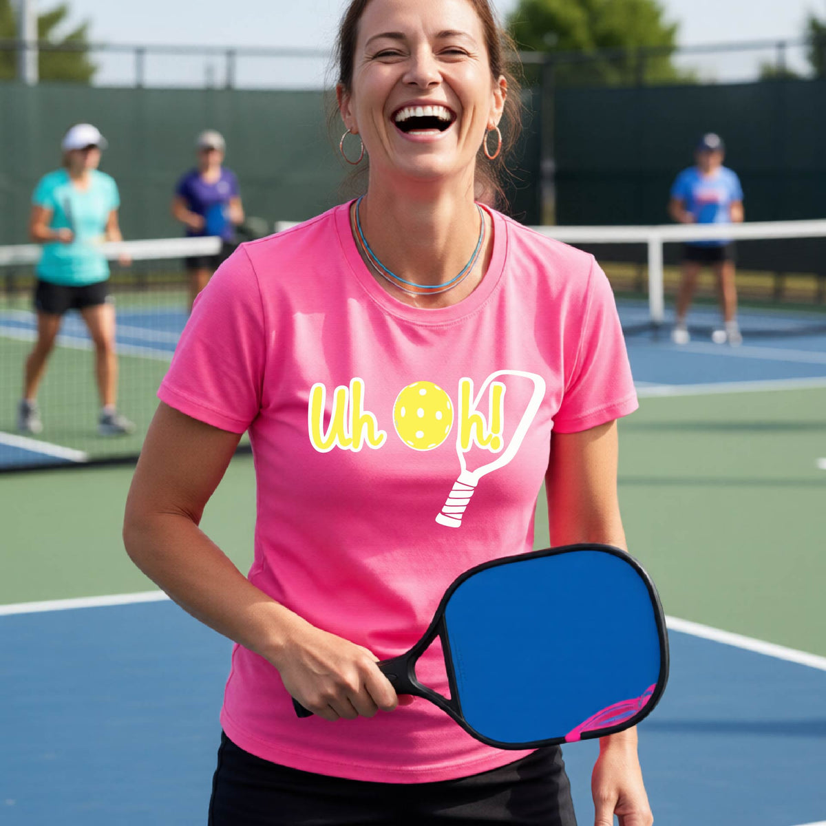 A woman on a pickleball court wearing a pink short-sleeve t-shirt with a white and yellow graphic of the words “Uh Oh!” with a yellow pickleball replacing the letter O on the front of the shirt, over the top of a pickleball paddle.