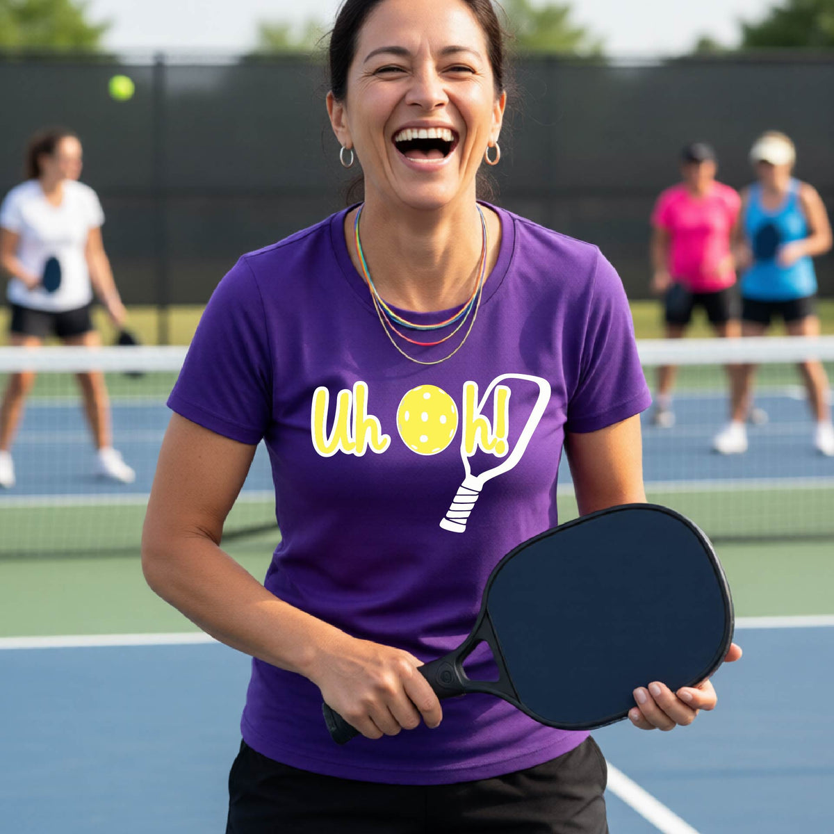 A woman on a pickleball court wearing a purple short-sleeve t-shirt with a white and yellow graphic of the words “Uh Oh!” with a yellow pickleball replacing the letter O on the front of the shirt, over the top of a pickleball paddle.