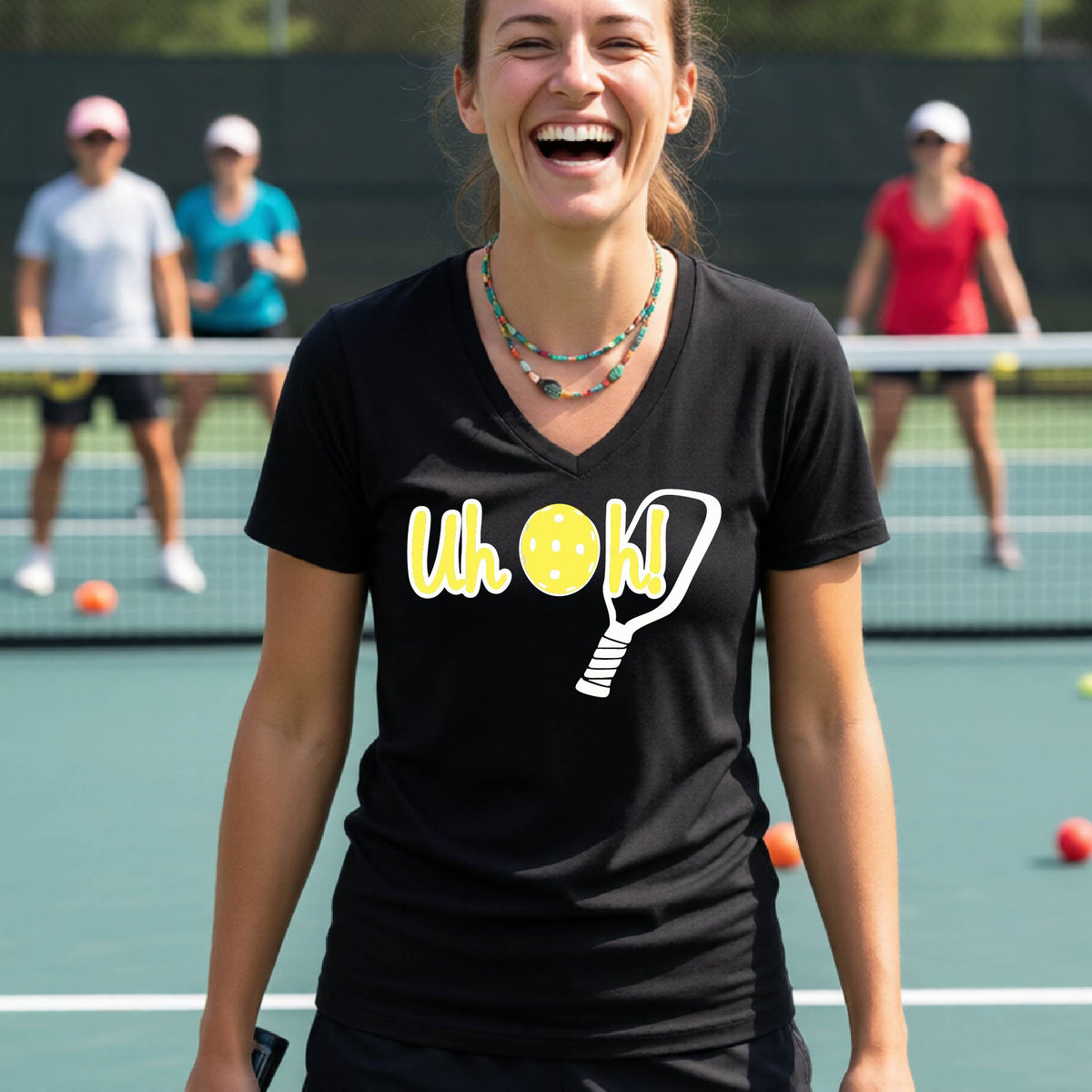 A woman on a pickleball court wearing a black  short-sleeve v-neck t-shirt with a white and yellow graphic of the words “Uh Oh!” with a yellow pickleball replacing the letter O on the front of the shirt, over the top of a pickleball paddle.