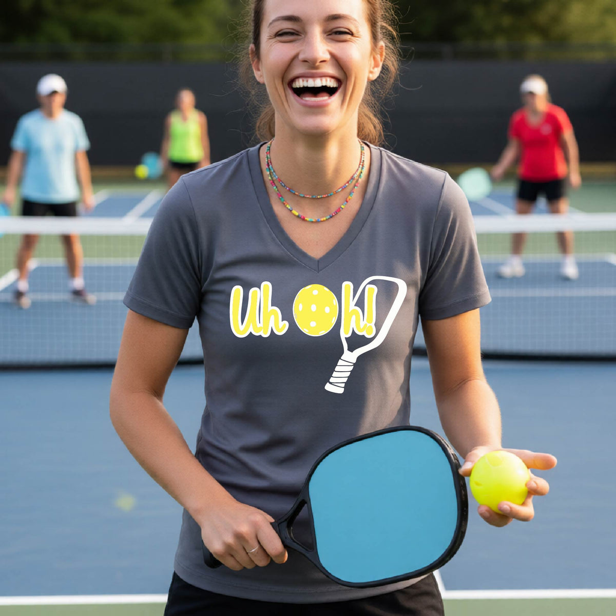 A woman on a pickleball court wearing a gray short-sleeve v-neck t-shirt with a white and yellow graphic of the words “Uh Oh!” with a yellow pickleball replacing the letter O on the front of the shirt, over the top of a pickleball paddle.