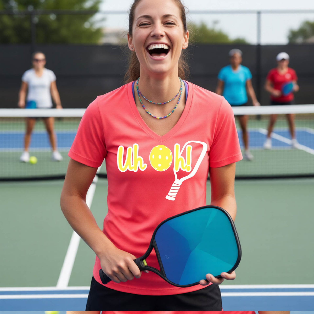 A woman on a pickleball court wearing a coral pink short-sleeve v-neck t-shirt with a white and yellow graphic of the words “Uh Oh!” with a yellow pickleball replacing the letter O on the front of the shirt, over the top of a pickleball paddle.