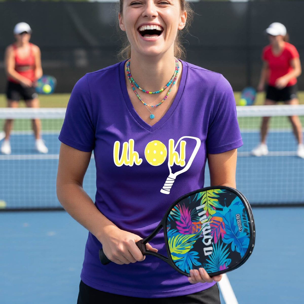 A woman on a pickleball court wearing a purple short-sleeve v-neck t-shirt with a white and yellow graphic of the words “Uh Oh!” with a yellow pickleball replacing the letter O on the front of the shirt, over the top of a pickleball paddle.