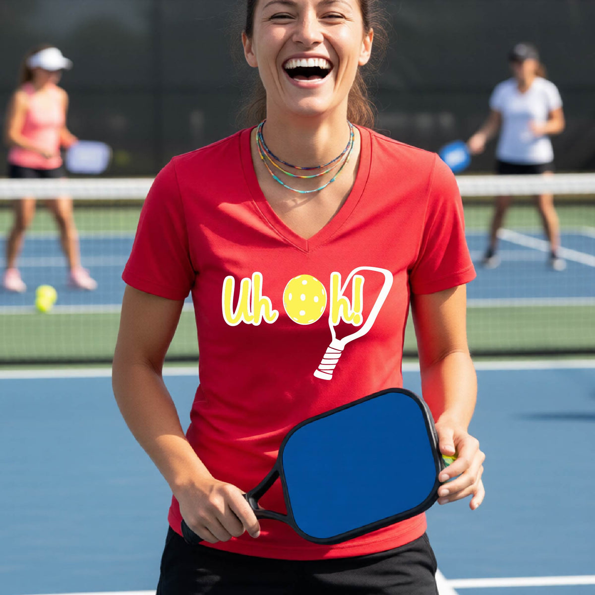 A woman on a pickleball court wearing a red short-sleeve v-neck t-shirt with a white and yellow graphic of the words “Uh Oh!” with a yellow pickleball replacing the letter O on the front of the shirt, over the top of a pickleball paddle.