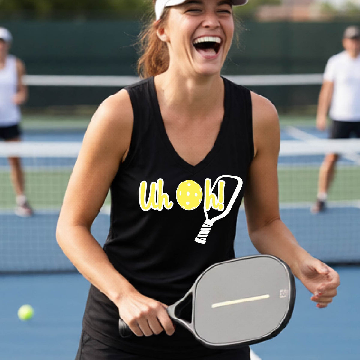 A woman on a pickleball court wearing a black short-sleeve v-neck t-shirt with a white and yellow graphic of the words “Uh Oh!” with a yellow pickleball replacing the letter O on the front of the shirt, over the top of a pickleball paddle.