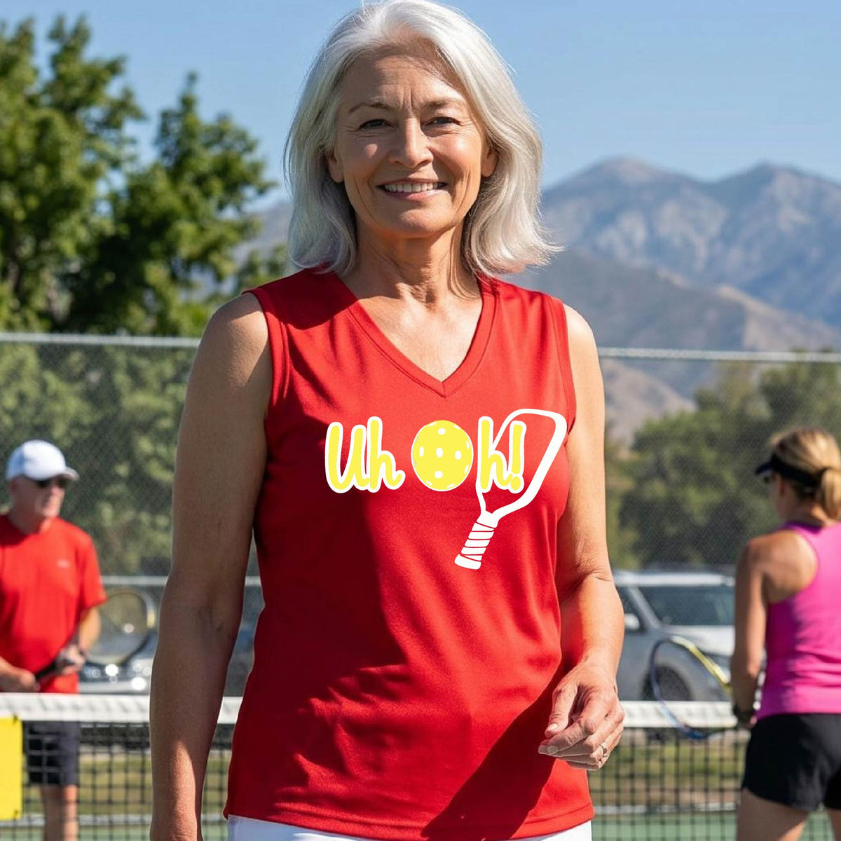 A woman on a pickleball court wearing a red short-sleeve v-neck t-shirt with a white and yellow graphic of the words “Uh Oh!” with a yellow pickleball replacing the letter O on the front of the shirt, over the top of a pickleball paddle.