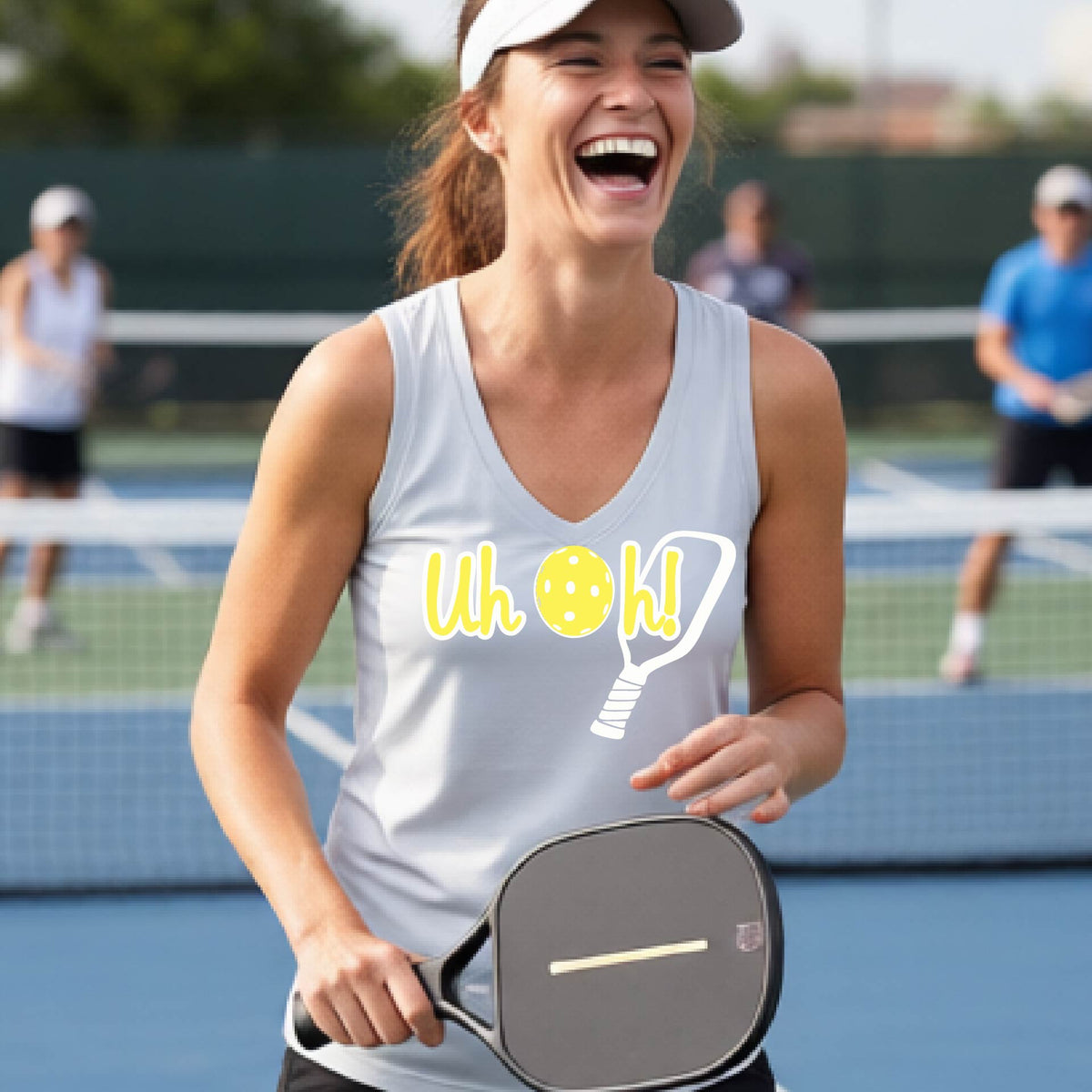A woman on a pickleball court wearing a silver gray short-sleeve v-neck t-shirt with a white and yellow graphic of the words “Uh Oh!” with a yellow pickleball replacing the letter O on the front of the shirt, over the top of a pickleball paddle.