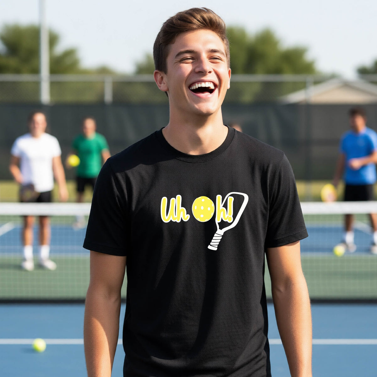 A young person on a pickleball court wearing a black short-sleeve t-shirt with a white and yellow graphic of the words “Uh Oh!” with a yellow pickleball replacing the letter O on the front of the shirt, over the top of a pickleball paddle.