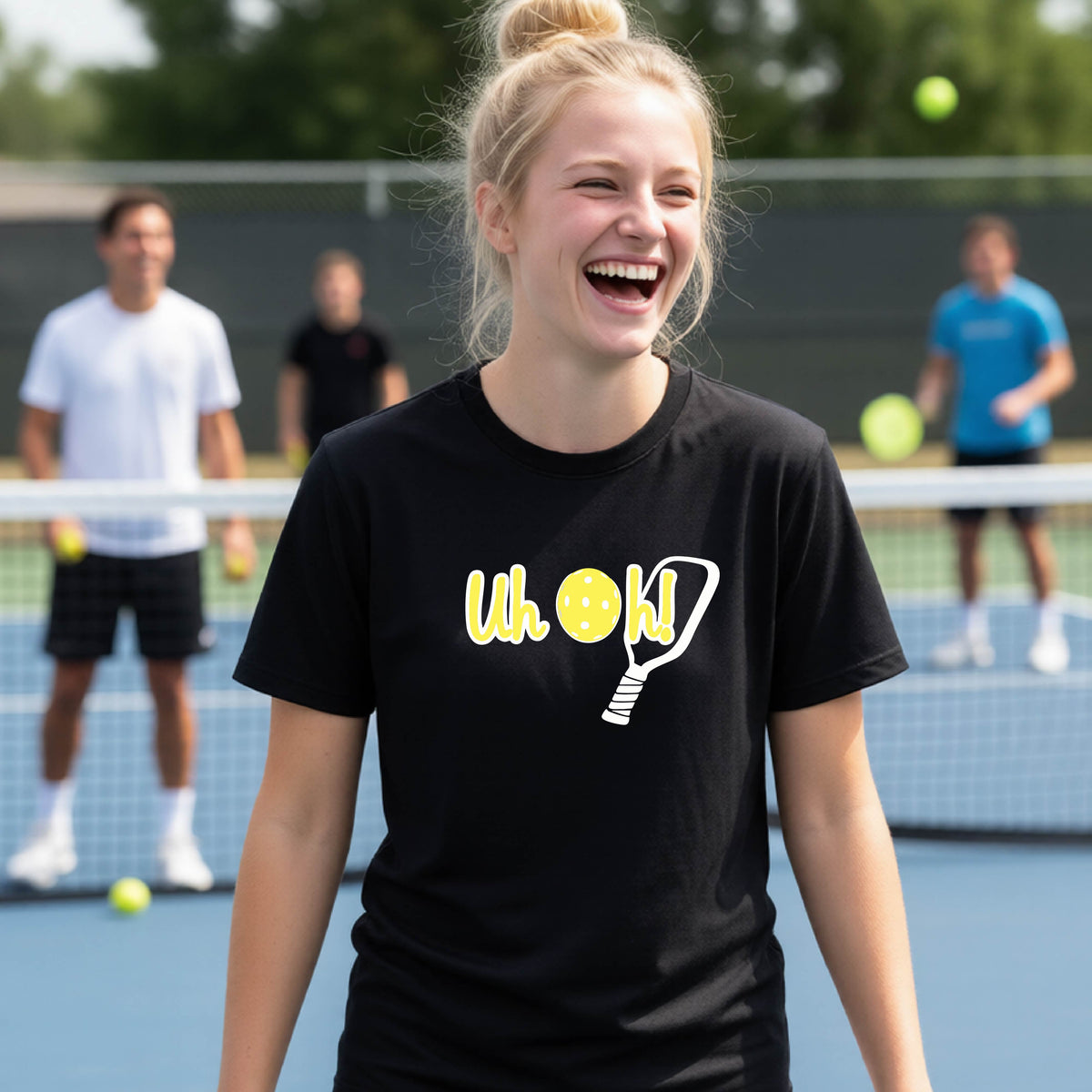 A young person on a pickleball court wearing a black short-sleeve t-shirt with a white and yellow graphic of the words “Uh Oh!” with a yellow pickleball replacing the letter O on the front of the shirt, over the top of a pickleball paddle.