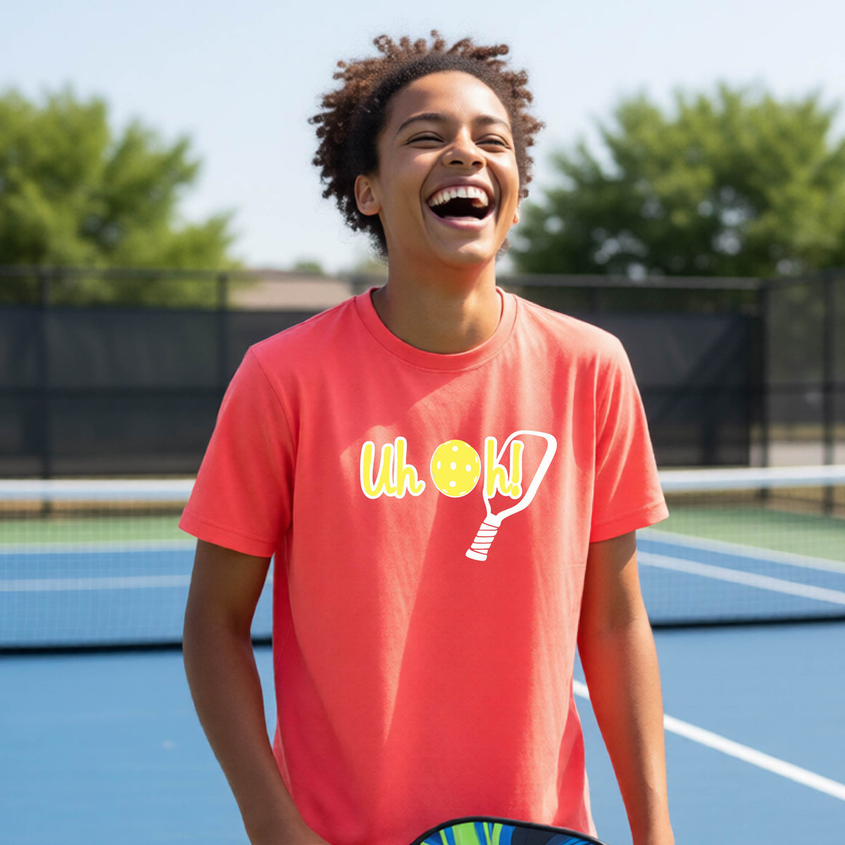 A young person on a pickleball court wearing a coral pink short-sleeve t-shirt with a white and yellow graphic of the words “Uh Oh!” with a yellow pickleball replacing the letter O on the front of the shirt, over the top of a pickleball paddle.