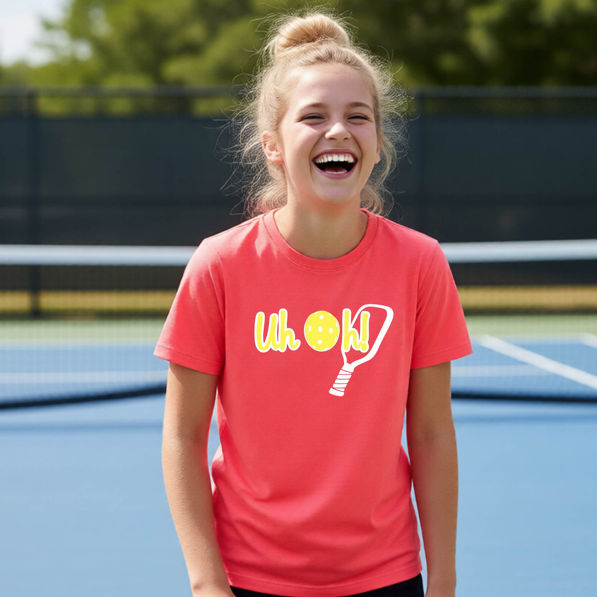 A young person on a pickleball court wearing a coral pink short-sleeve t-shirt with a white and yellow graphic of the words “Uh Oh!” with a yellow pickleball replacing the letter O on the front of the shirt, over the top of a pickleball paddle.