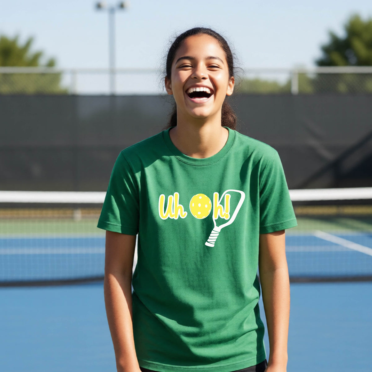 A young person on a pickleball court wearing a green short-sleeve t-shirt with a white and yellow graphic of the words “Uh Oh!” with a yellow pickleball replacing the letter O on the front of the shirt, over the top of a pickleball paddle.